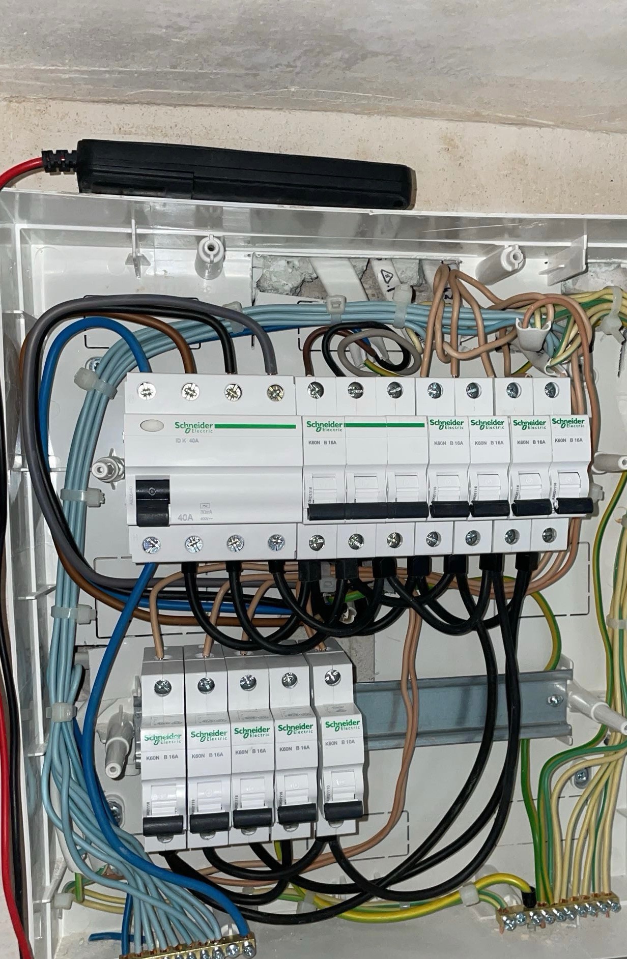 Rozdzielnia elektryczna Schneider Electric z bezpiecznikami i przewodami w różnych kolorach, zamontowana w białej obudowie. Widoczne ślady montażu na ścianie.