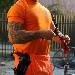 Na Ziher Silesia Shine - Mężczyzna w pomarańczowym stroju roboczym czyści ściągaczkę do okien czerwoną szmatką. Widoczny sprzęt do mycia okien i tatuaże na ramionach.