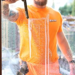 Na Ziher Silesia Shine - Mężczyzna w pomarańczowym stroju roboczym myje okno za pomocą ściągaczki, pozostawiając smugi wody. Widoczne drzewa w tle.