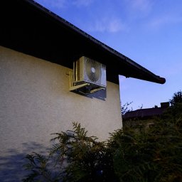 VentoTech - Biała jednostka zewnętrzna klimatyzacji AUX zamontowana na elewacji domu, widoczna z dołu na tle wieczornego nieba i zieleni. Ujęcie z niskiego kąta.