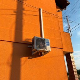 VentoTech - Zewnętrzna jednostka klimatyzatora Haier zamontowana na pomarańczowej elewacji budynku, z białą rurą maskującą instalację. Widoczny cień słupa i kabli, rynna oraz antena na dachu.