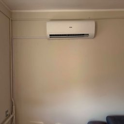 VentoTech - Biała jednostka klimatyzacyjna marki Haier zamontowana na jasnobeżowej ścianie w pomieszczeniu, z widocznym białym korytem instalacyjnym biegnącym wzdłuż ściany i w dół, obok fragmentu ciemnej sofy.