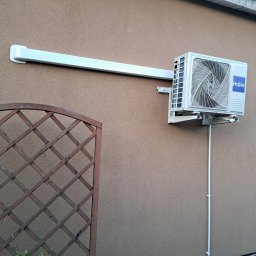 VentoTech - Zainstalowana zewnętrzna jednostka klimatyzacji Haier na teksturowanej, jasnobrązowej ścianie, z białym korytem instalacyjnym i fragmentem drewnianej kratki ogrodowej.