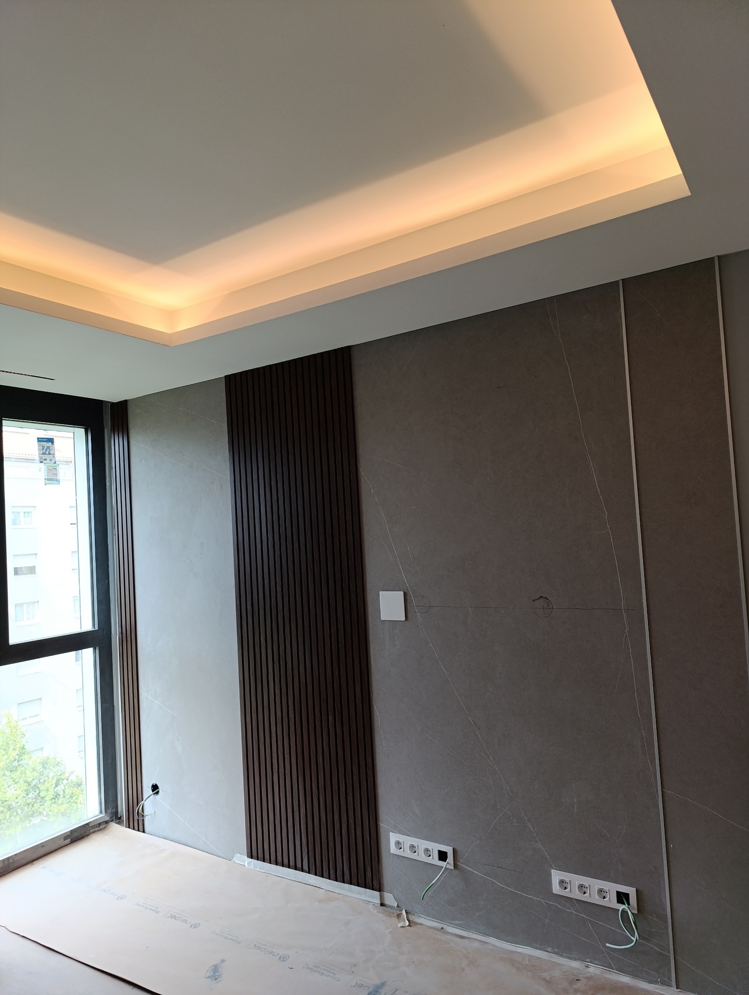 Wykończone ściany w pokoju z panelami dekoracyjnymi i płytami imitującymi marmur. Widoczne gniazdka elektryczne i podświetlany sufit. Podłoga zabezpieczona folią.