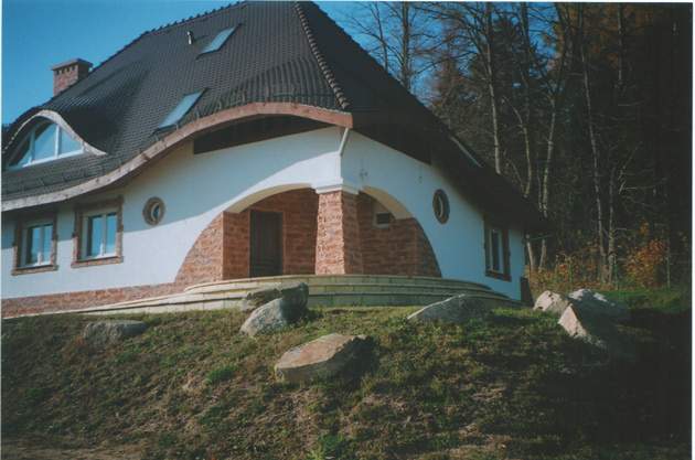 Nietypowy budynek jednorodzinny z falistym dachem krytym dachówką, białymi ścianami i kamiennym wykończeniem elewacji, okrągłe okno, schody wejściowe z kamienia, otoczenie z zielonym trawnikiem...