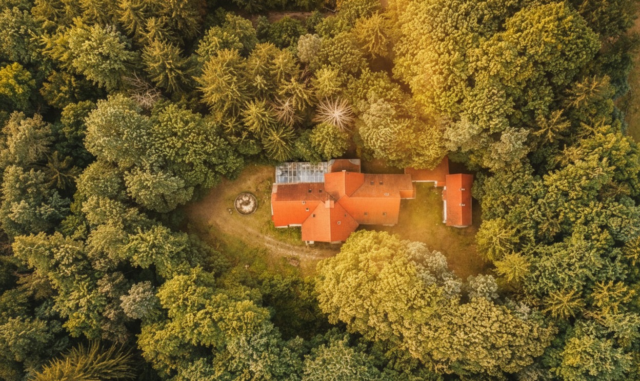 Widok z drona na budynek z czerwoną dachówką otoczony lasem. Ujęcie z góry, ciepłe światło słoneczne. Na podwórku widoczna studnia.