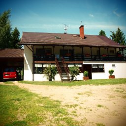 Biebrza24 - Dwupiętrowy budynek z balkonem i schodami zewnętrznymi, czerwony samochód w garażu, trawnik i drzewa w tle, błękitne niebo. Widok z przodu, słoneczny dzień.