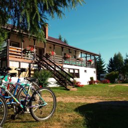 Biebrza24 - Pensjonat w Sztabinie z balkonem udekorowanym kwiatami. Na pierwszym planie rowery, w tle zieleń i błękitne niebo. Słoneczny dzień.
