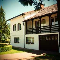 Biebrza24 - Dwukondygnacyjny dom z balkonem i garażem, biała elewacja, brązowe okna i dachówka. Uporządkowany ogród z niskim żywopłotem i drzewami. Słoneczny dzień.