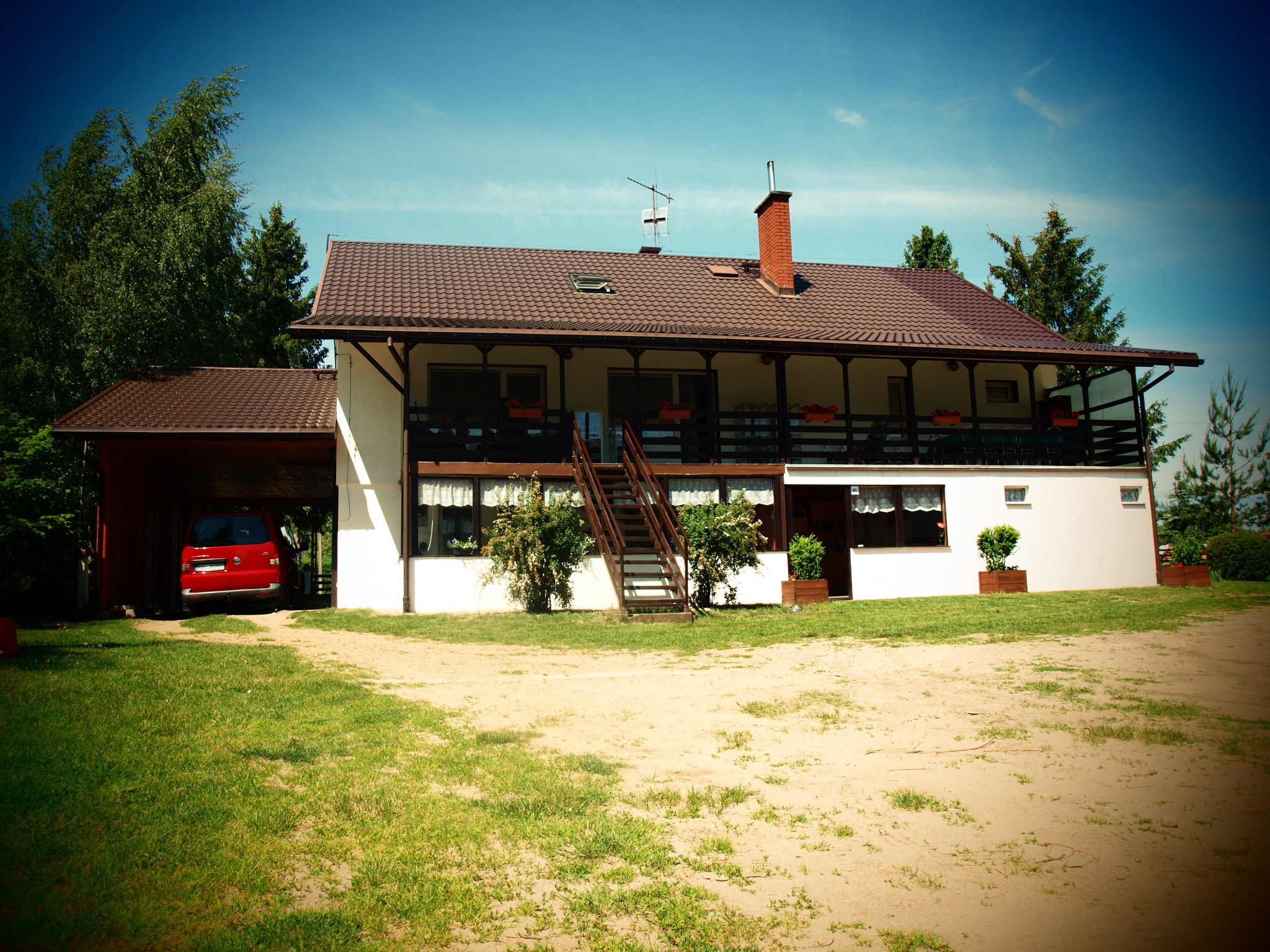 Dwupiętrowy budynek z balkonem i schodami zewnętrznymi, czerwony samochód w garażu, trawnik i drzewa w tle, błękitne niebo. Widok z przodu, słoneczny dzień.