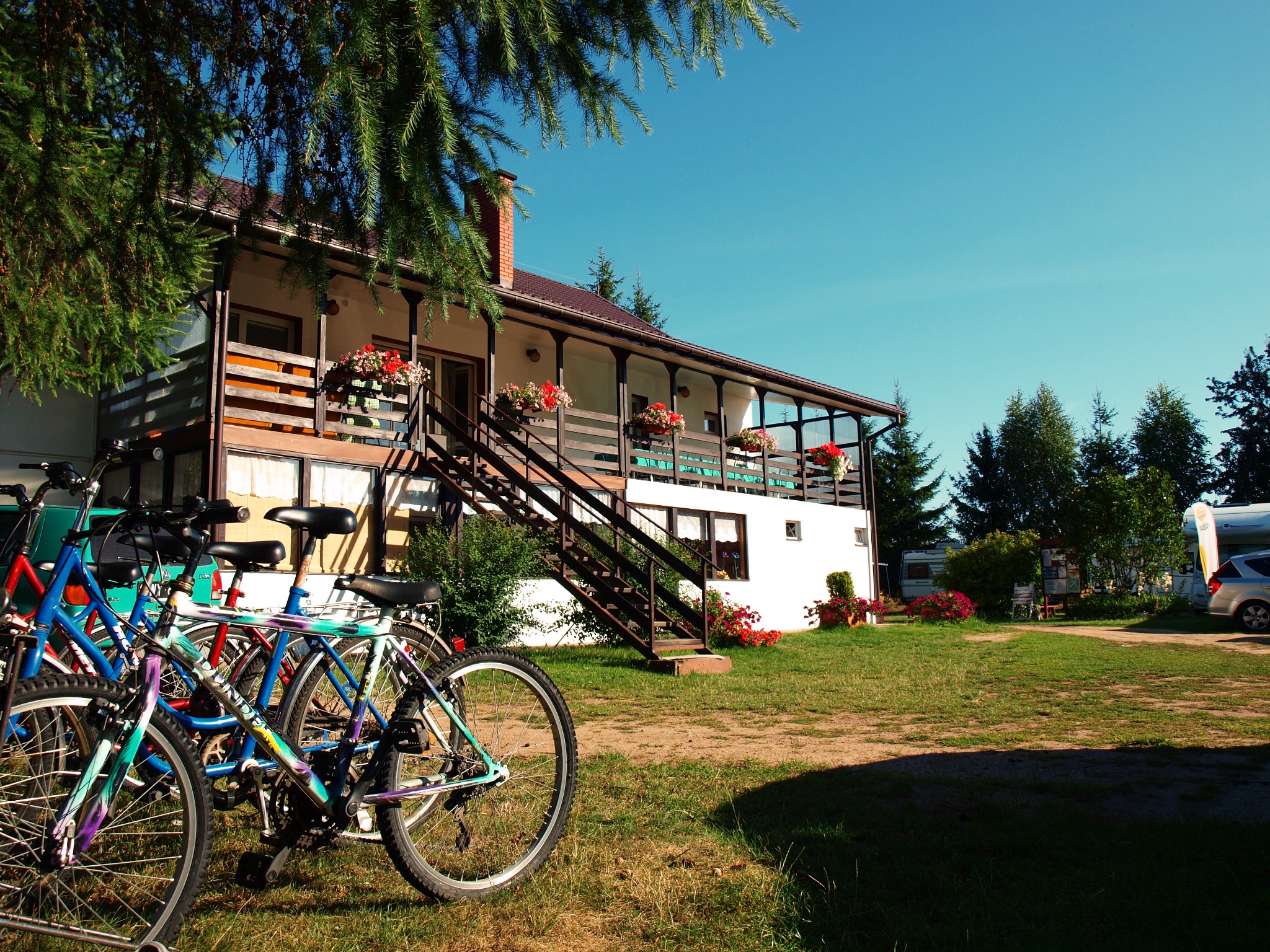 Pensjonat w Sztabinie z balkonem udekorowanym kwiatami. Na pierwszym planie rowery, w tle zieleń i błękitne niebo. Słoneczny dzień.