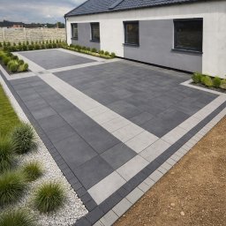 RADLUK - Nowoczesny podjazd z kostki betonowej w odcieniach szarości, z dekoracyjnymi pasami i obrzeżami. Widok na dom z szarą dachówką i starannie zagospodarowany ogród.