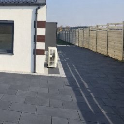 RADLUK - Szara kostka brukowa na podjeździe domu jednorodzinnego, z widocznym fragmentem elewacji i ogrodzeniem w tle. Długi cień rzucany na kostkę. Słoneczny dzień.