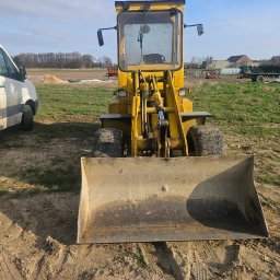 RADLUK - Żółta ładowarka na polu, widok z przodu, z widocznymi śladami opon na ziemi i stertą piasku w tle. Obok zaparkowany biały bus.
