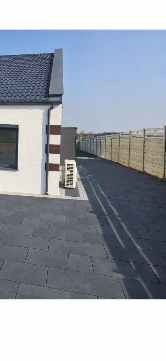 Szara kostka brukowa na podjeździe domu jednorodzinnego, z widocznym fragmentem elewacji i ogrodzeniem w tle. Długi cień rzucany na kostkę. Słoneczny dzień.