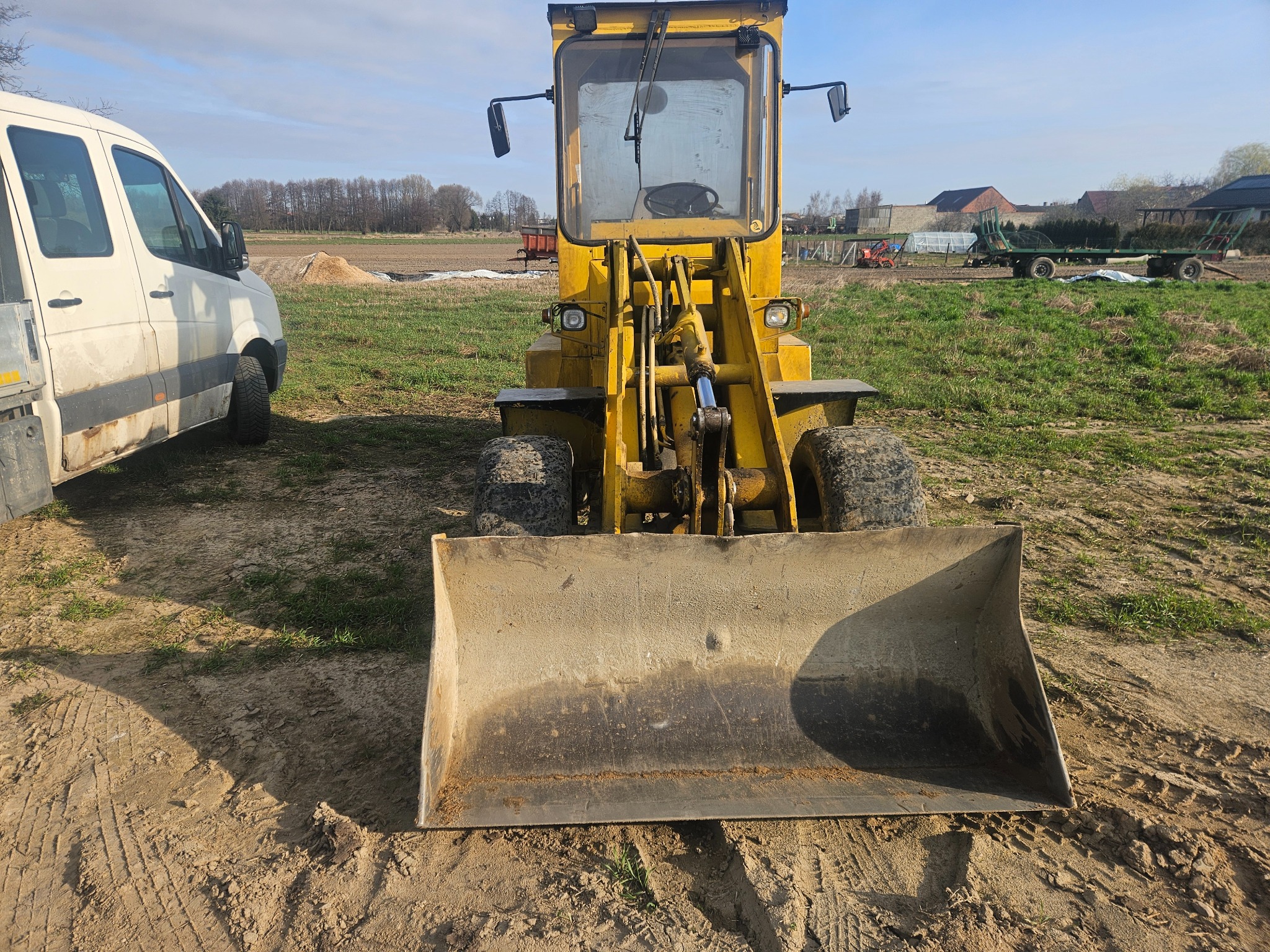 Żółta ładowarka na polu, widok z przodu, z widocznymi śladami opon na ziemi i stertą piasku w tle. Obok zaparkowany biały bus.