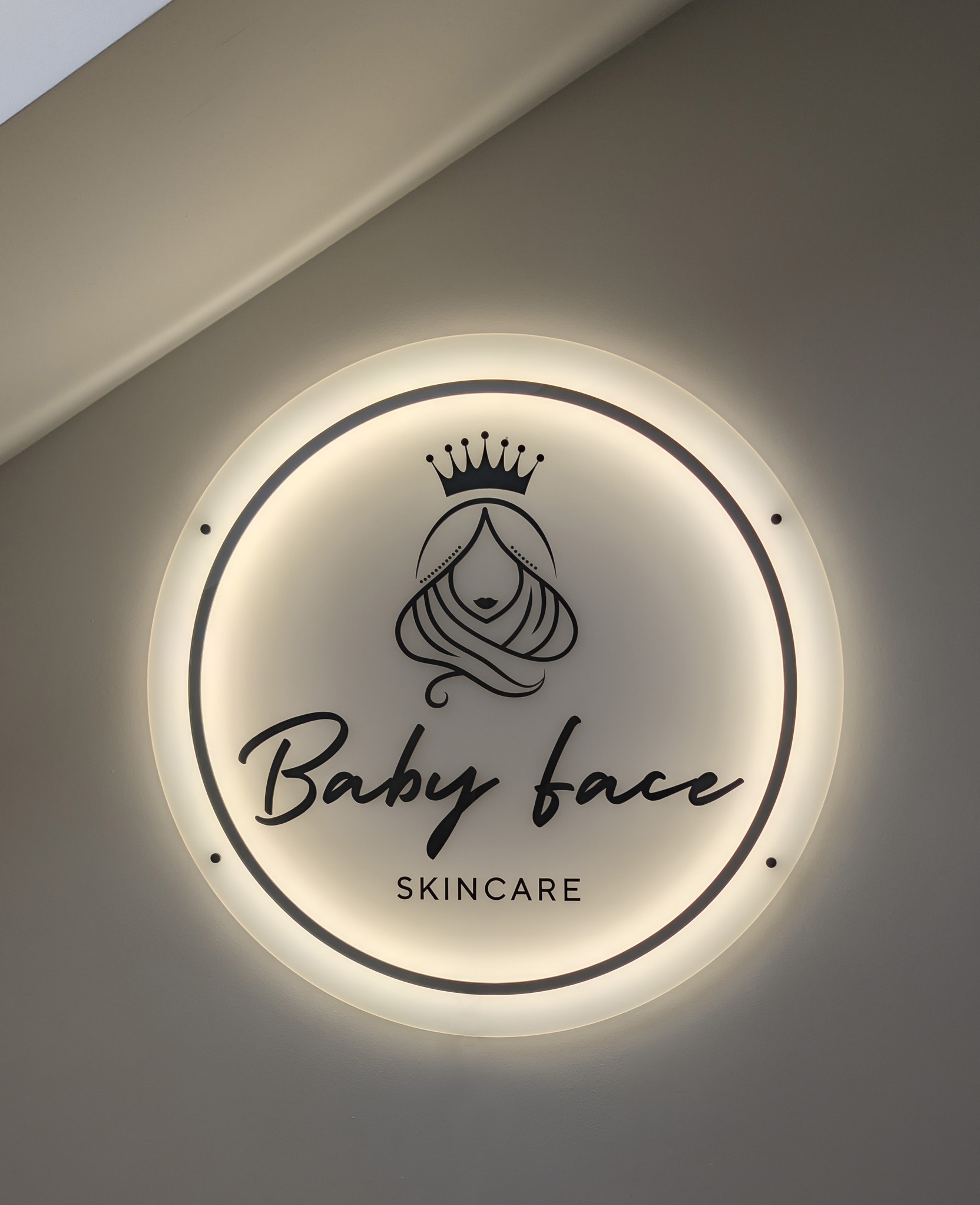 Podświetlane, okrągłe logo 'Baby face SKINCARE' z minimalistyczną grafiką kobiety w koronie, zamontowane na szarej ścianie, oświetlone od tyłu.