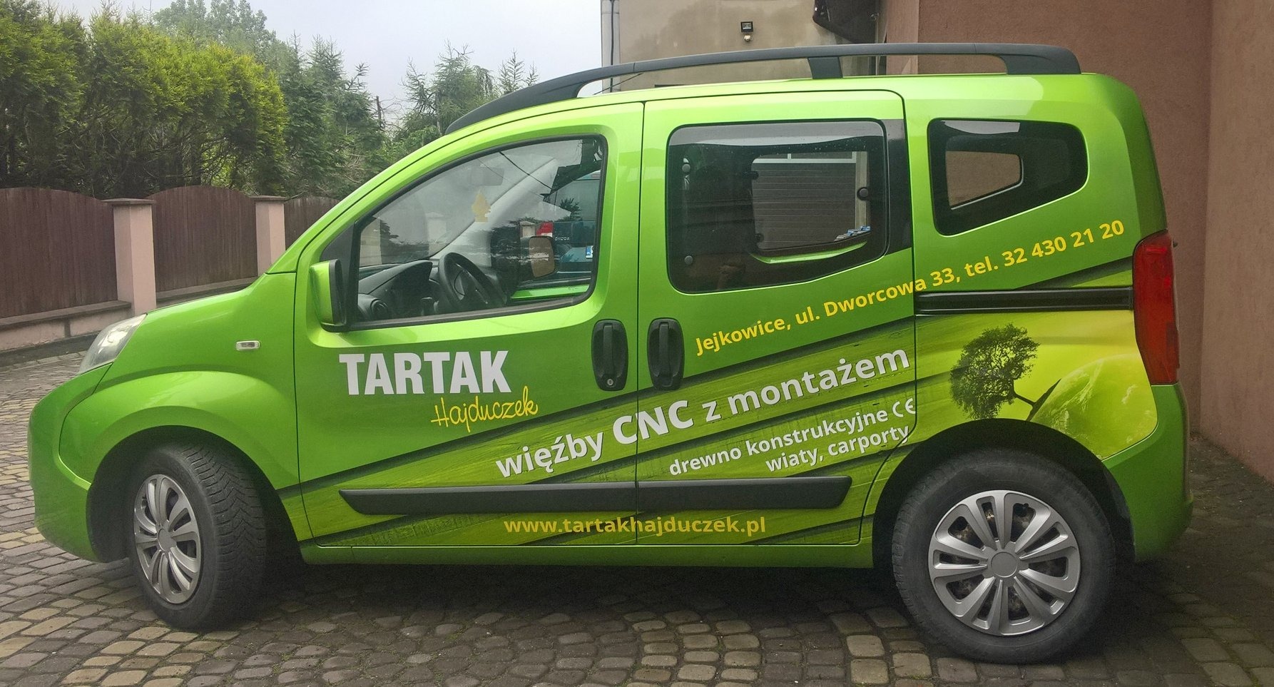 Zielony samochód dostawczy z grafiką reklamową tartaku, prezentujący usługi CNC, więźby i konstrukcje drewniane. Widoczne dane kontaktowe i adres strony internetowej.