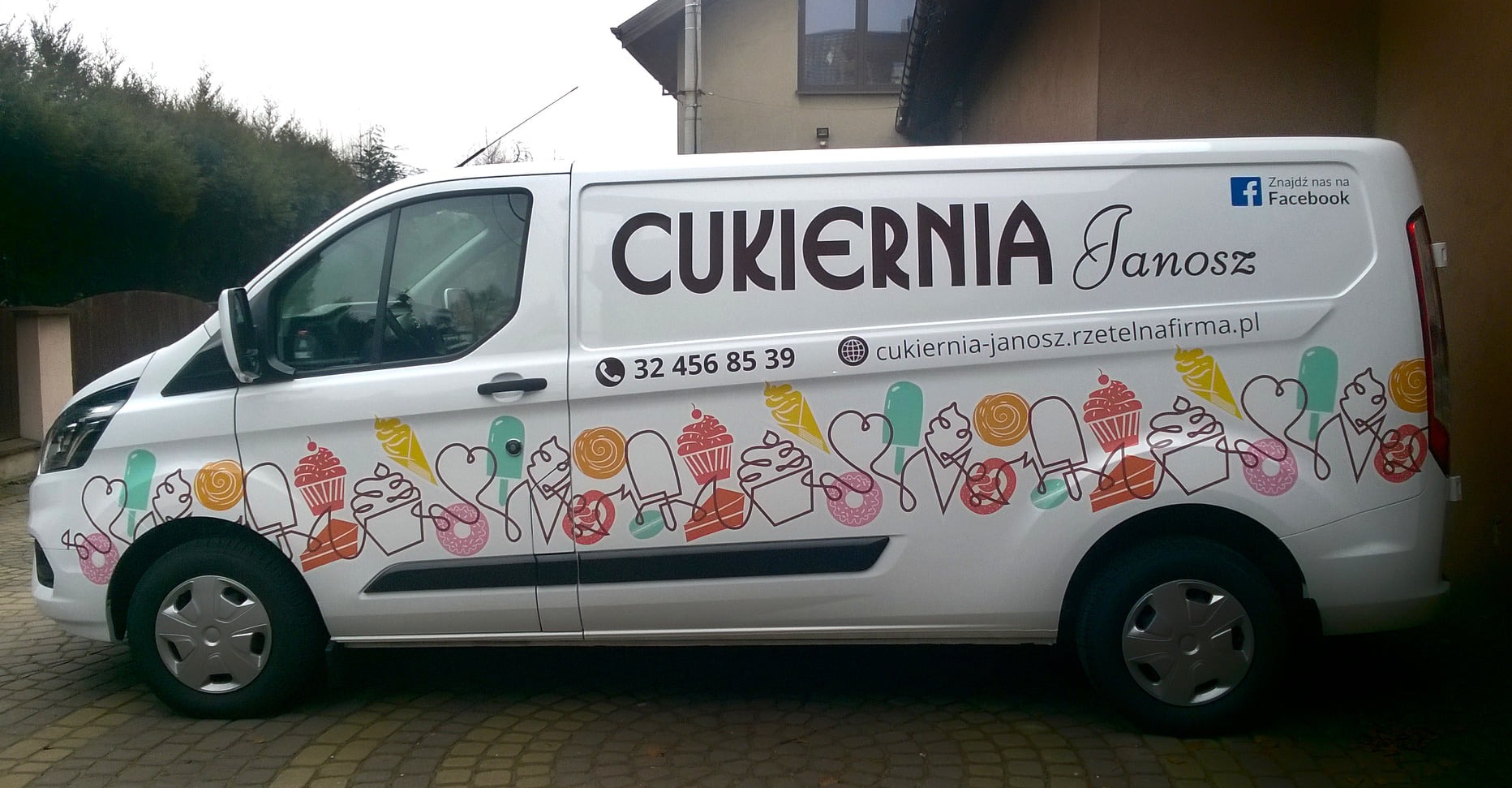 Biały van z grafiką cukierniczą i danymi kontaktowymi 'Cukiernia Janosz', oklejony na całej długości, zaparkowany na brukowanej nawierzchni.