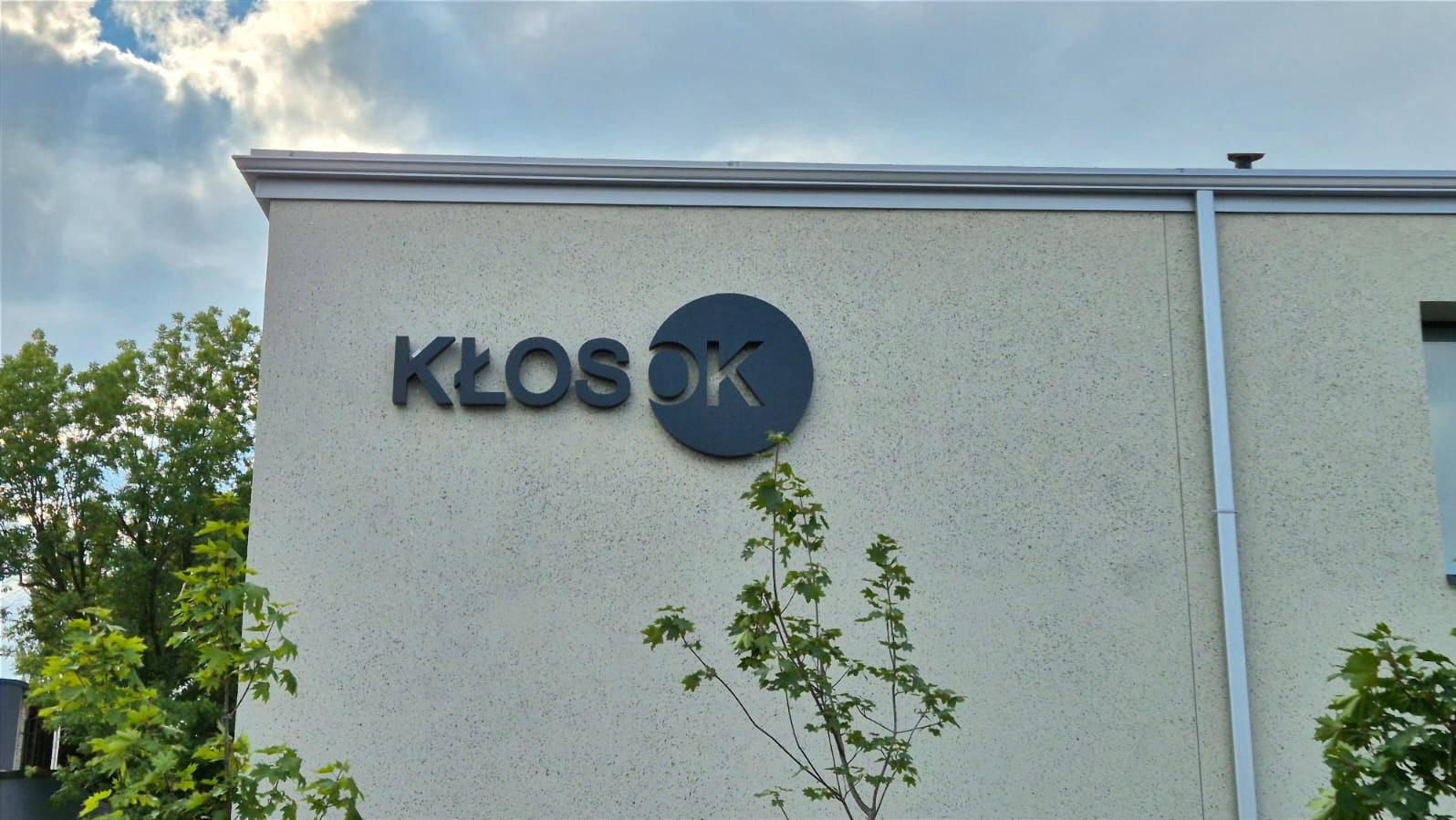 Elewacja budynku z napisem 'KŁOS OK' w ciemnoszarym kolorze, otoczona zielenią drzew. Ujęcie z dołu, widoczne fragmenty nieba z chmurami.