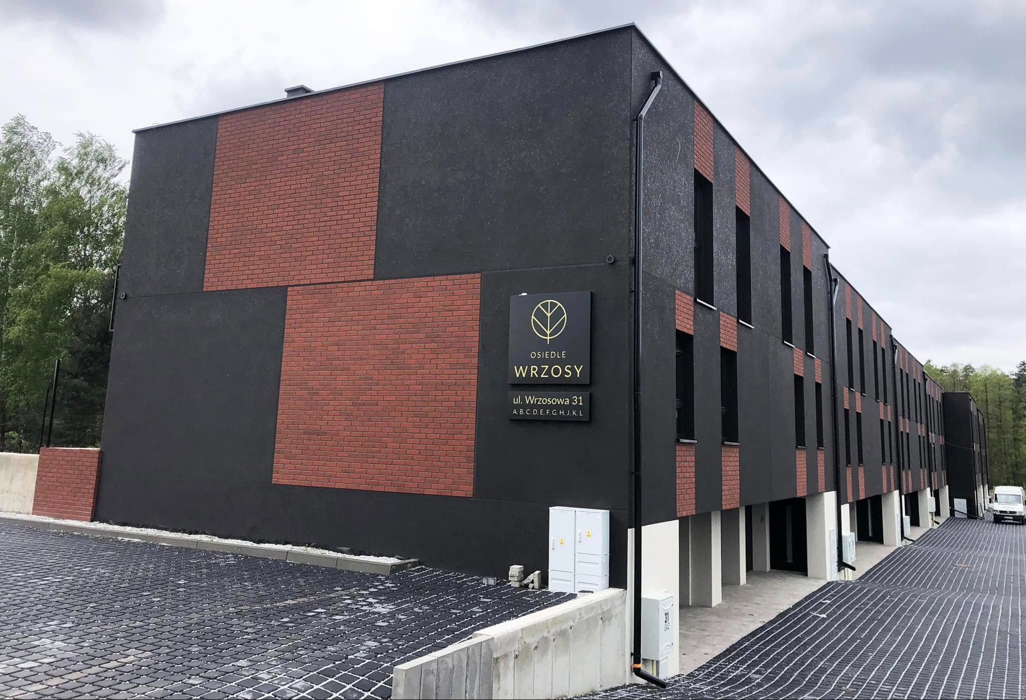 Nowoczesny budynek osiedla Wrzosy z czarną elewacją i ceglanymi akcentami, widoczny z perspektywy parkingu z ażurową nawierzchnią. Minimalistyczny design.