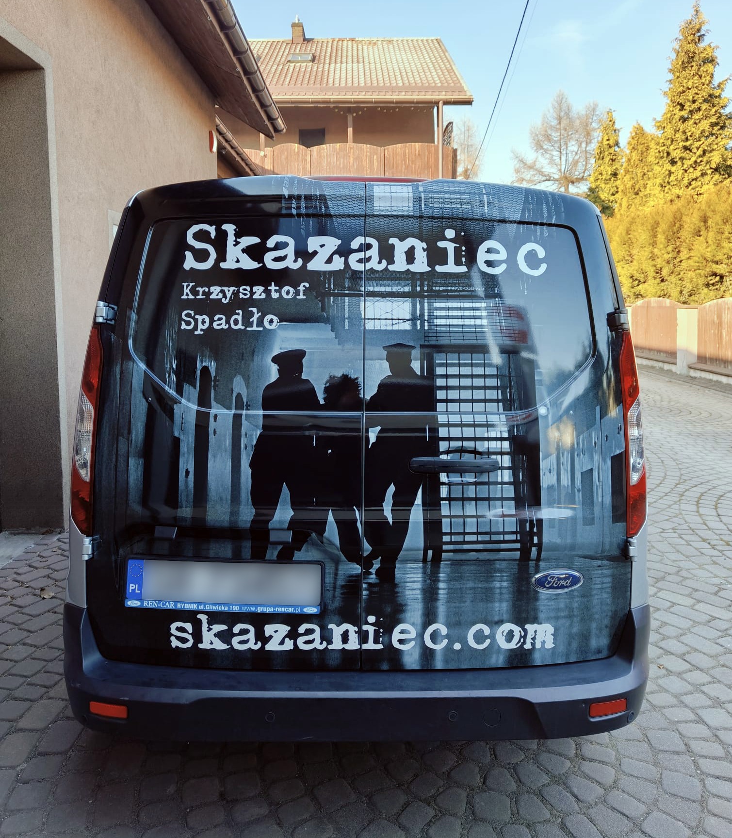 Oklejony tył srebrnego Forda Transita z czarno-białą grafiką filmową, napisem 'Skazaniec' i adresem strony internetowej. Widoczne tablice rejestracyjne i logo Forda.