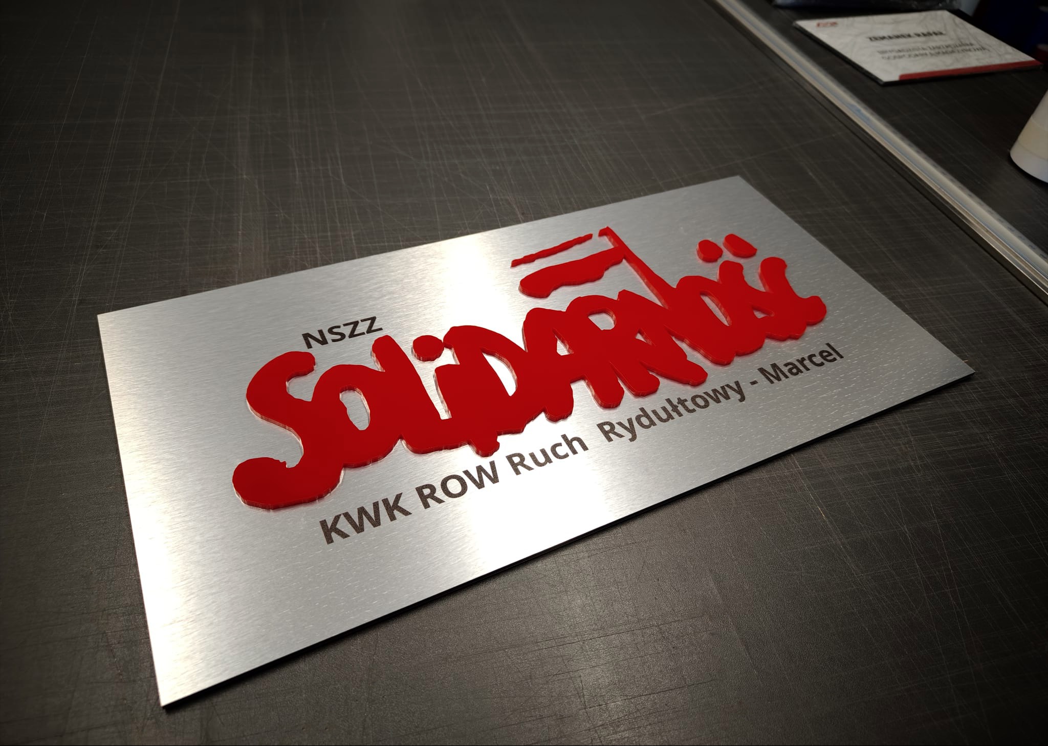 Metalowa tabliczka z czerwonym, wypukłym napisem 'Solidarność' i tekstem 'KWK ROW Ruch Rydułtowy - Marcel' na szarym, teksturowanym tle.