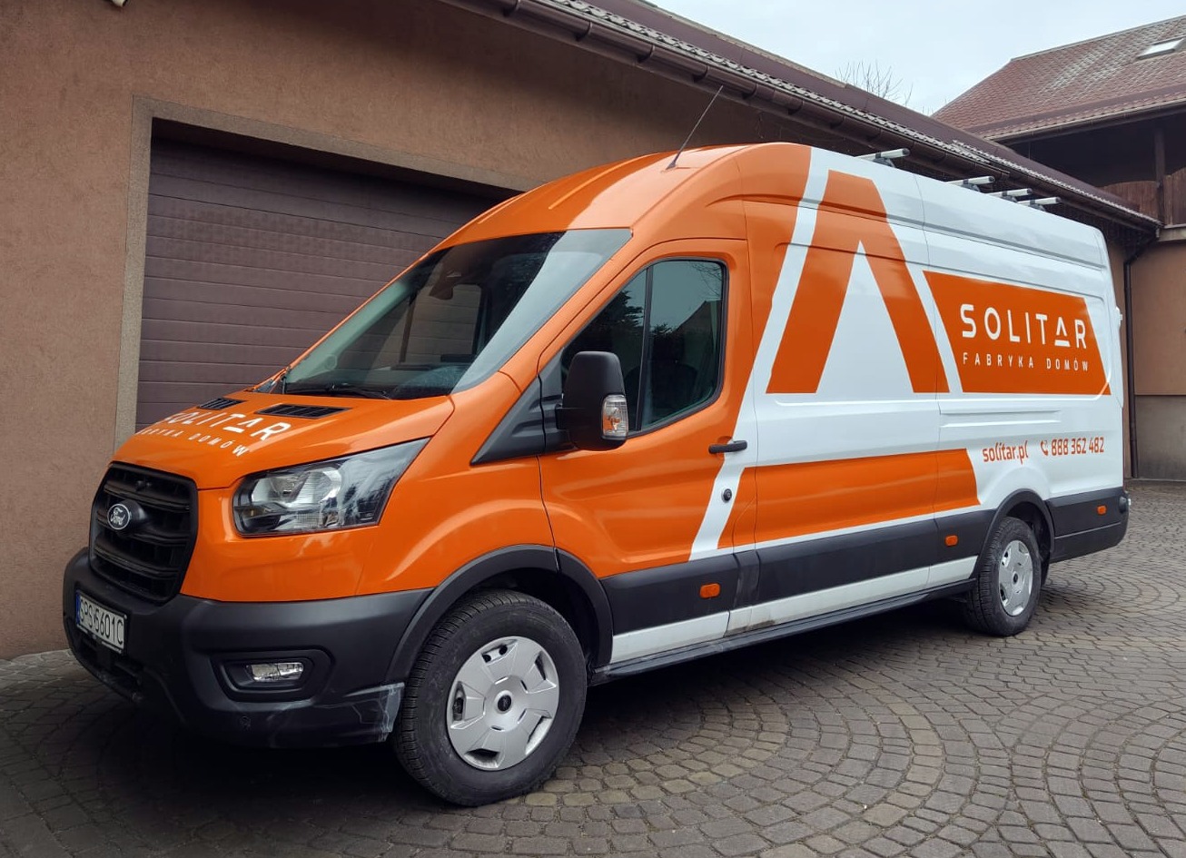 Pomarańczowo-biały van Ford Transit z logo 'Solitar Fabryka Domów' na tle budynku z brązową bramą garażową i kostki brukowej. Widok z boku, lekki skos.