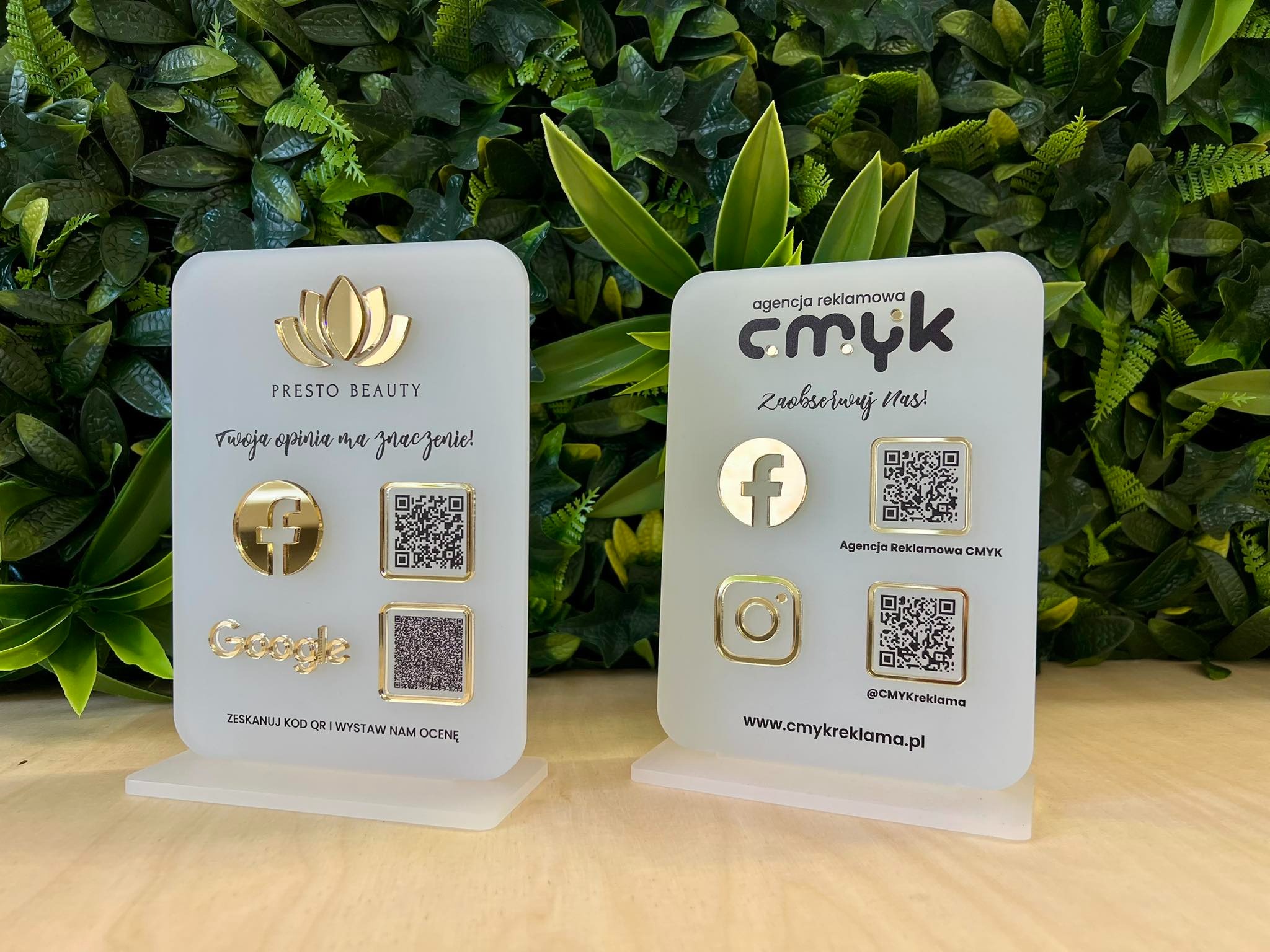 Dwa stojaki reklamowe z logo firm i kodami QR na tle zielonej ściany roślinnej. Stojaki z akrylu z elementami w kolorze złotym. Prezentacja usług reklamowych.