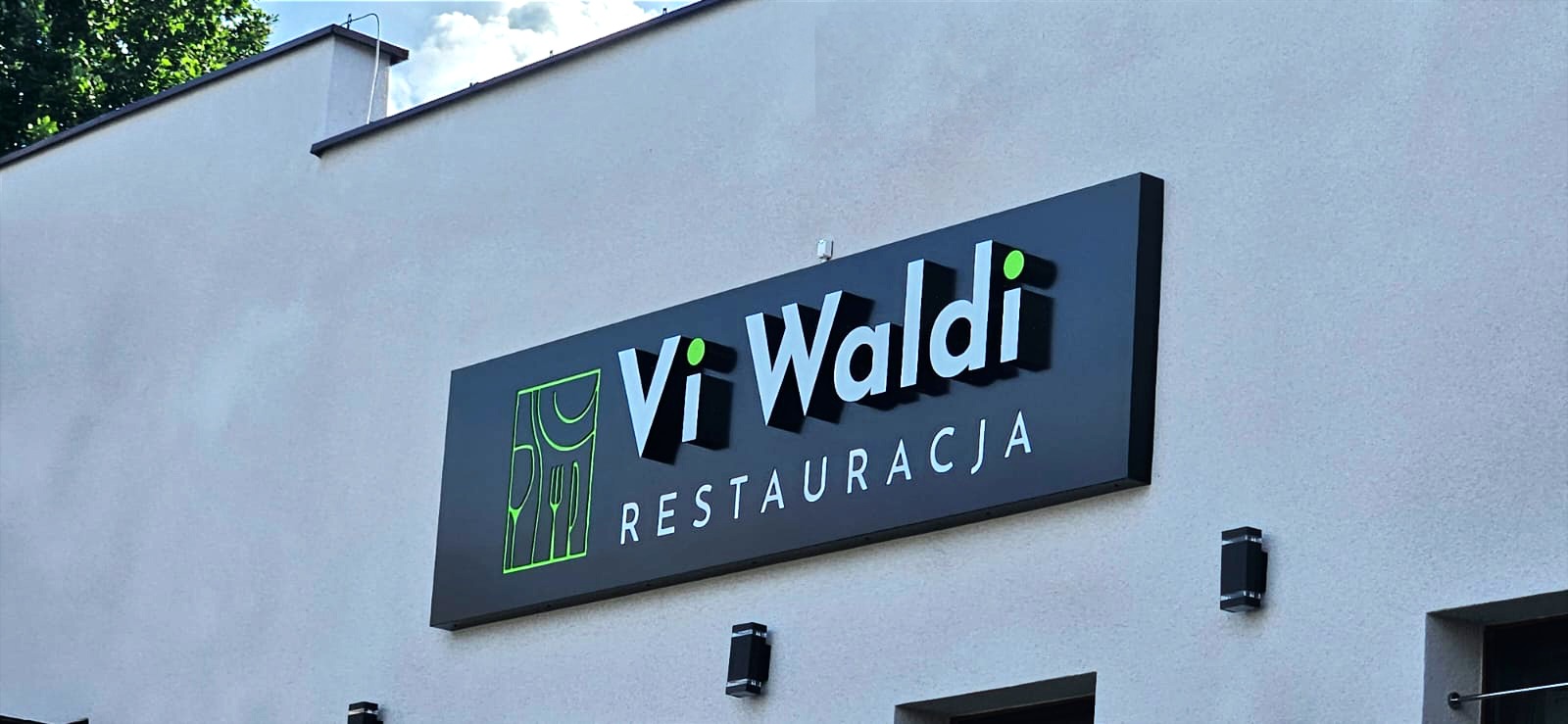 Elegancki szyld restauracji Vi Waldi z logo i nazwą na czarnym tle, zamontowany na jasnej elewacji budynku. Widoczne oświetlenie zewnętrzne.