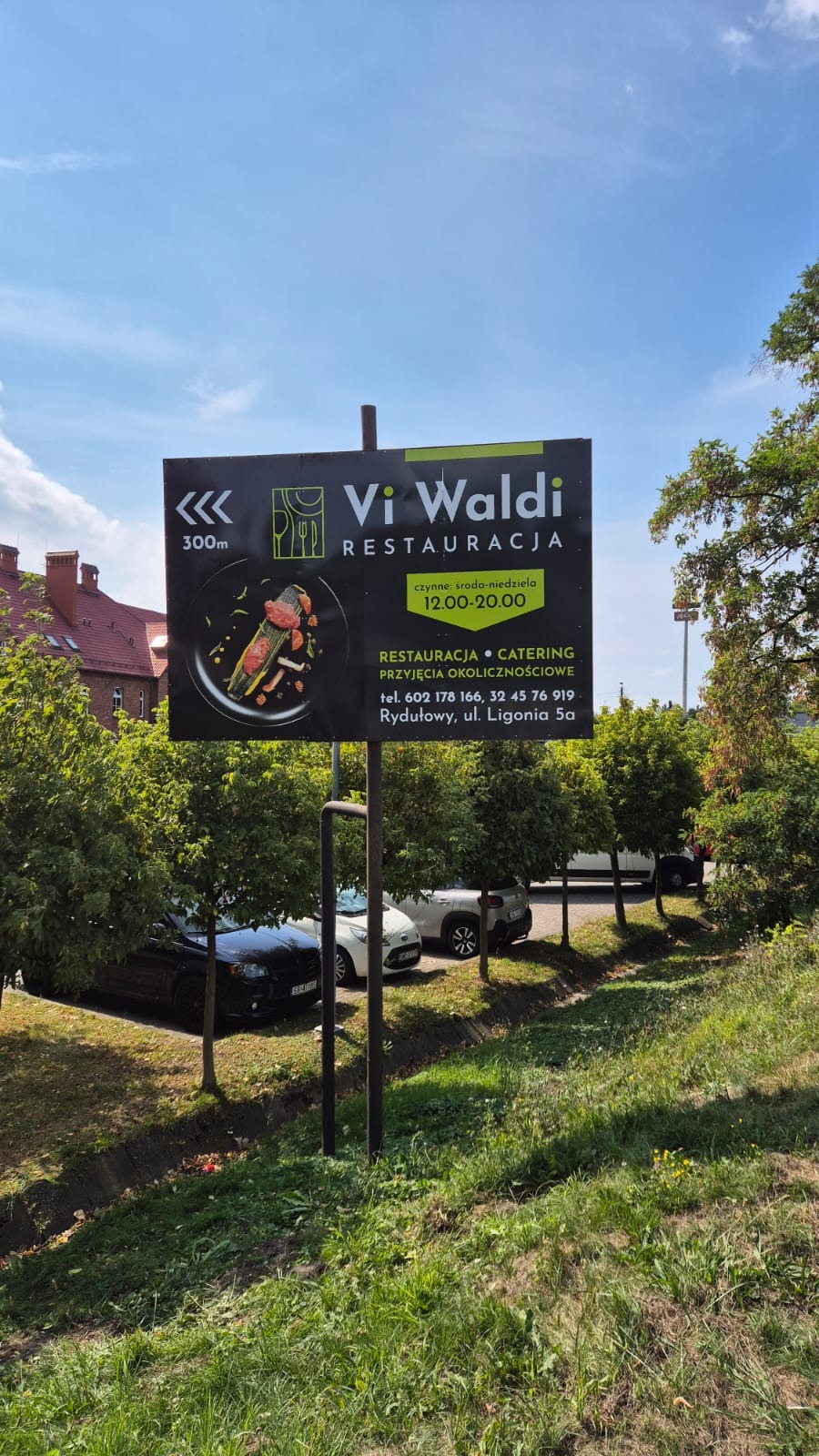 Billboard restauracji Vi Waldi z daniem na czarnym talerzu, godziny otwarcia i dane kontaktowe na tle błękitnego nieba i zieleni drzew. Reklama lokalu gastronomicznego.
