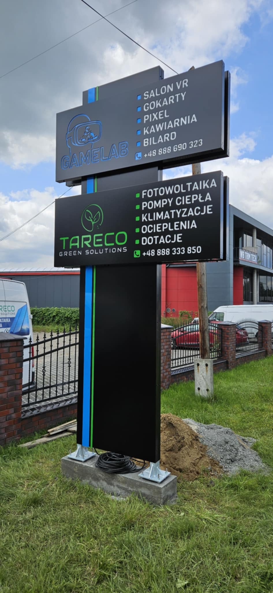 Dwustronny pylon reklamowy z logo firm Gamelab i Tareco Green Solutions, z listą usług i numerami telefonów, umieszczony na zewnątrz budynku. Widoczne elementy montażowe.