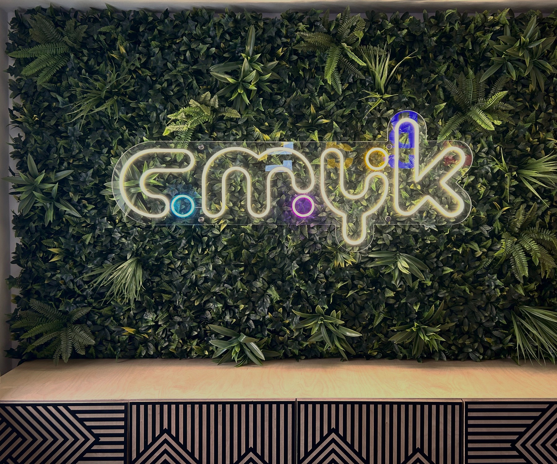 Neonowy napis 'cmylk' na tle ściany z zielonych roślin, umieszczony nad szafką z geometrycznym wzorem. Oświetlenie tworzy unikalny efekt wizualny.