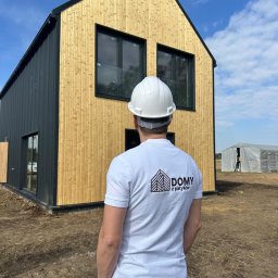 D.Z.P. SPÓŁKA Z OGRANICZONĄ ODPOWIEDZIALNOŚCIĄ - Dwukondygnacyjny dom szkieletowy z jasną elewacją drewnianą i ciemnymi panelami. Mężczyzna w kasku patrzy na budynek. W tle widoczny namiot.