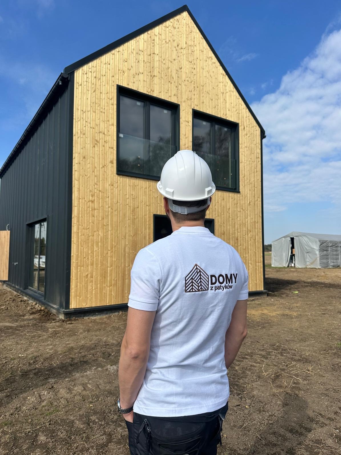 Dwukondygnacyjny dom szkieletowy z jasną elewacją drewnianą i ciemnymi panelami. Mężczyzna w kasku patrzy na budynek. W tle widoczny namiot.