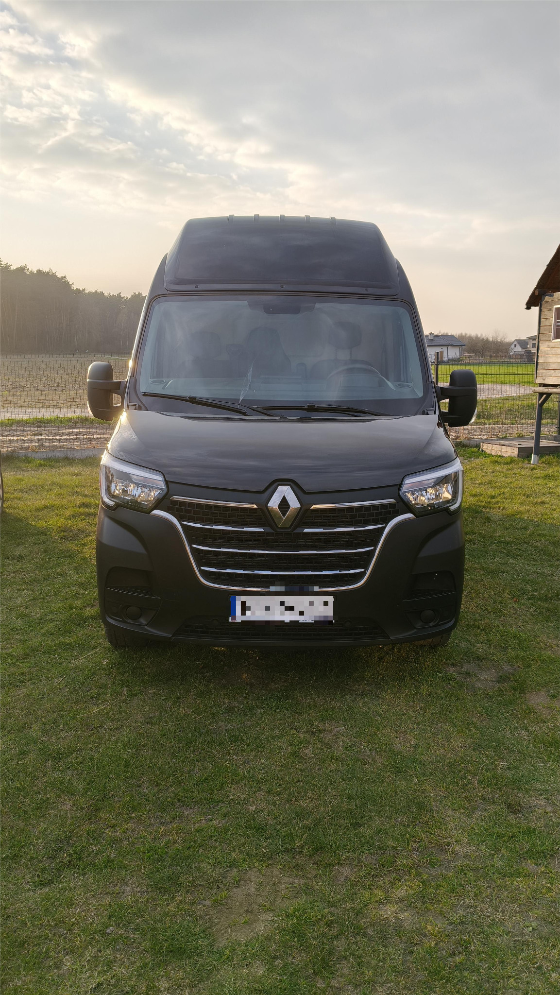Czarny Renault Master z wysokim dachem, zaparkowany na trawie, widok z przodu. Tablice rejestracyjne zamazane. W tle drzewa i budynek.