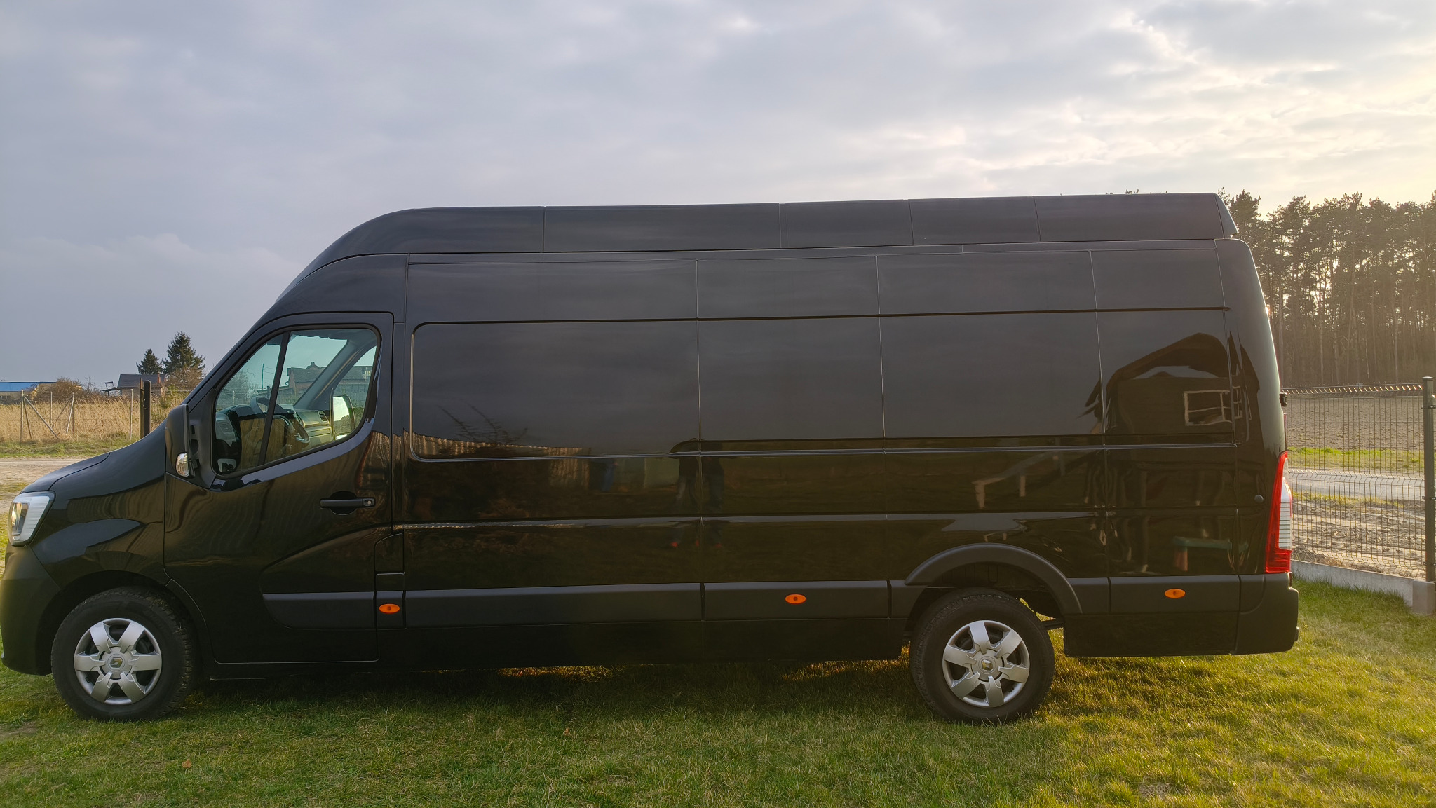 Czarny bus dostawczy Renault Master na trawie, widok z boku, z refleksami budynków w lakierze. Tło: drzewa i pochmurne niebo. Idealny do transportu i przeprowadzek.