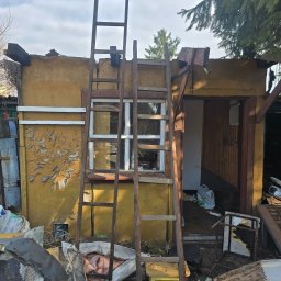 Gasiorex - Zniszczony, żółty budynek z odpadającą farbą, podparty drewnianą drabiną. Wokół gruz, śmieci i resztki mebli. Widoczne okno i otwarte wejście do budynku.