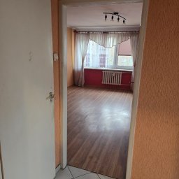 Gasiorex - Pusty pokój z drewnianą podłogą widziany przez otwarte drzwi. Pomarańczowe ściany, białe zasłony i grzejnik pod oknem. Widoczny fragment korytarza z białą podłogą.