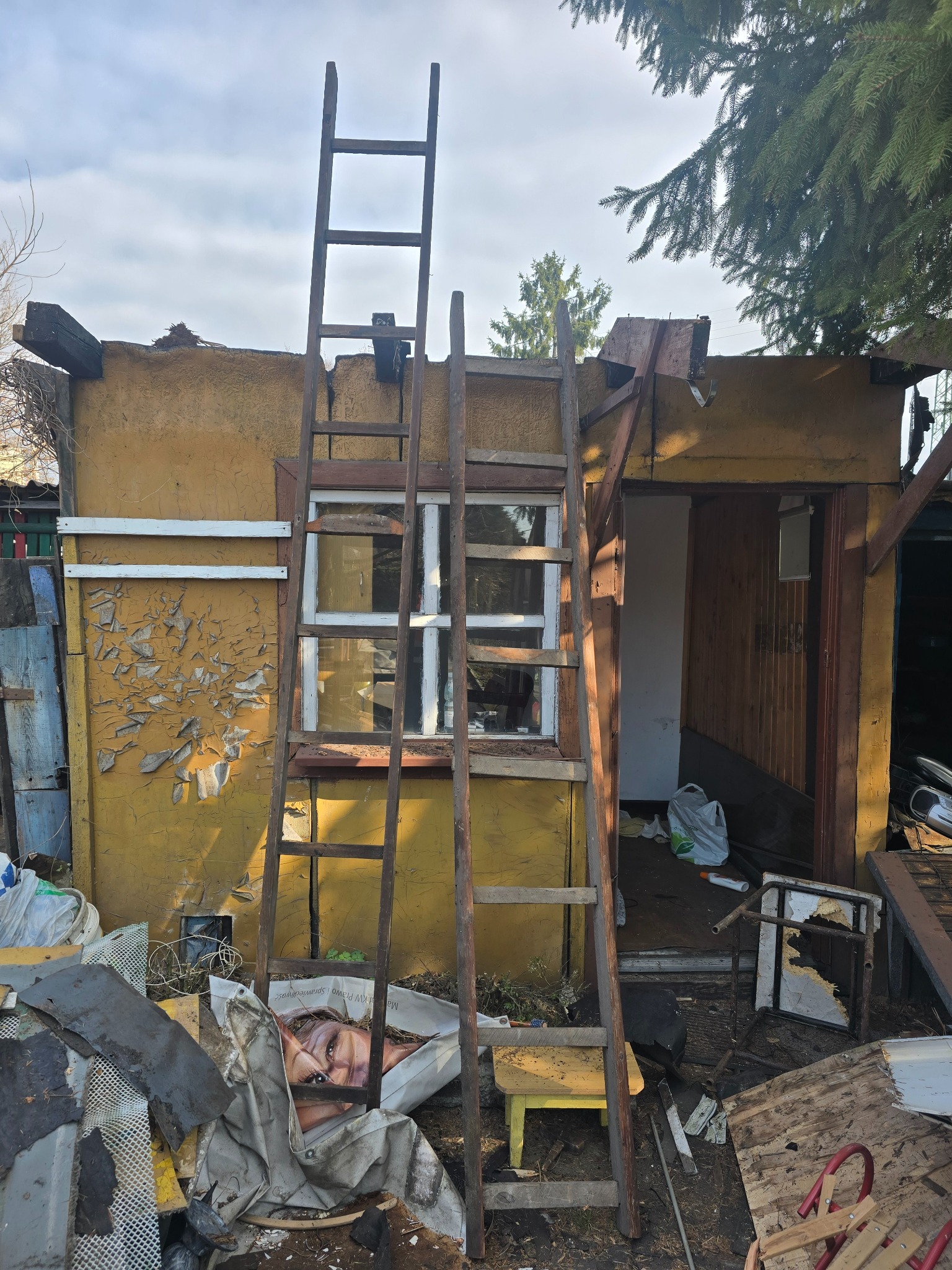 Zniszczony, żółty budynek z odpadającą farbą, podparty drewnianą drabiną. Wokół gruz, śmieci i resztki mebli. Widoczne okno i otwarte wejście do budynku.