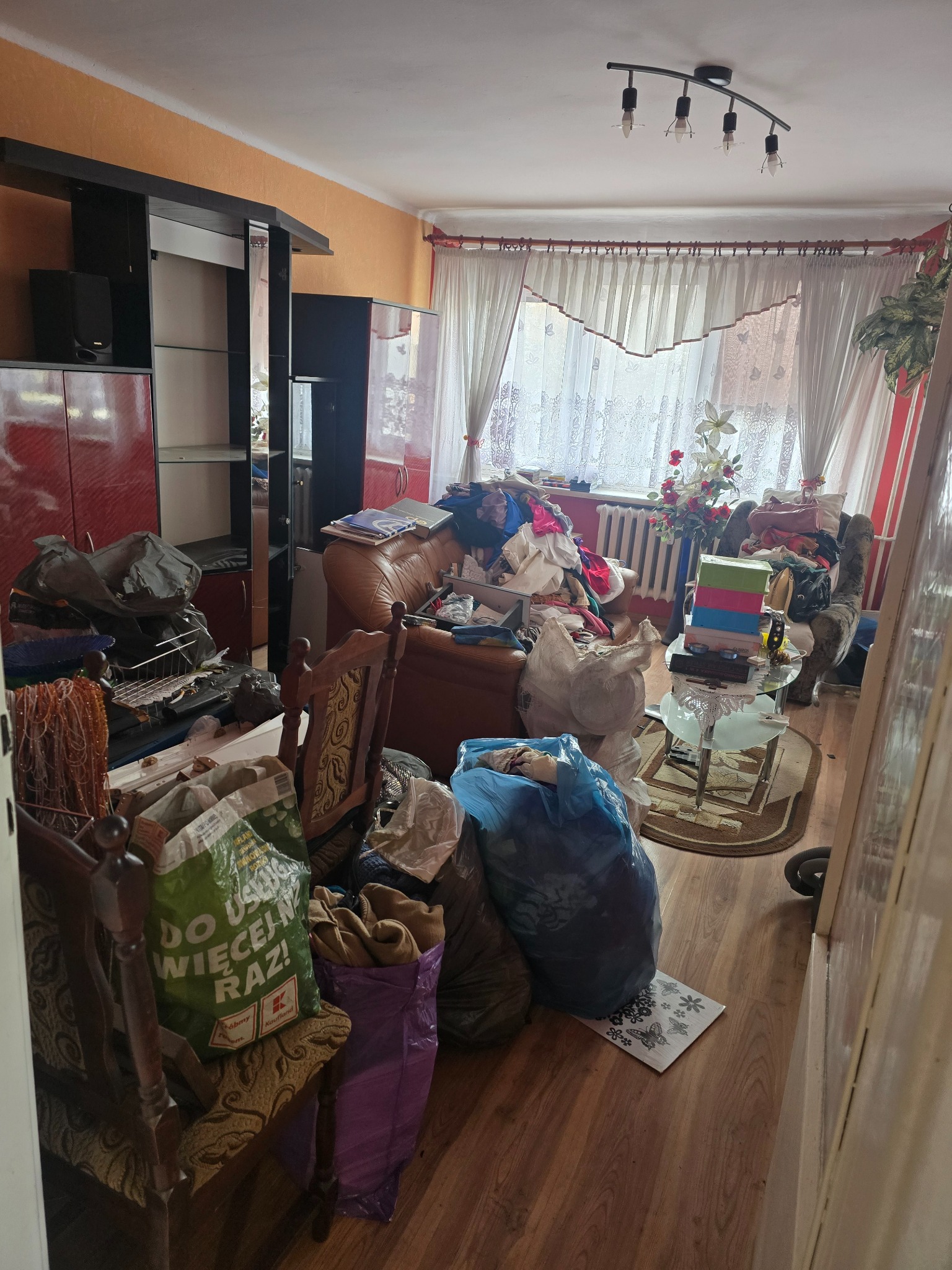 Zabrudzone mieszkanie z meblami i workami pełnymi śmieci. Widok na pokój z bałaganem, ukazujący potrzebę profesjonalnego opróżniania i sprzątania.