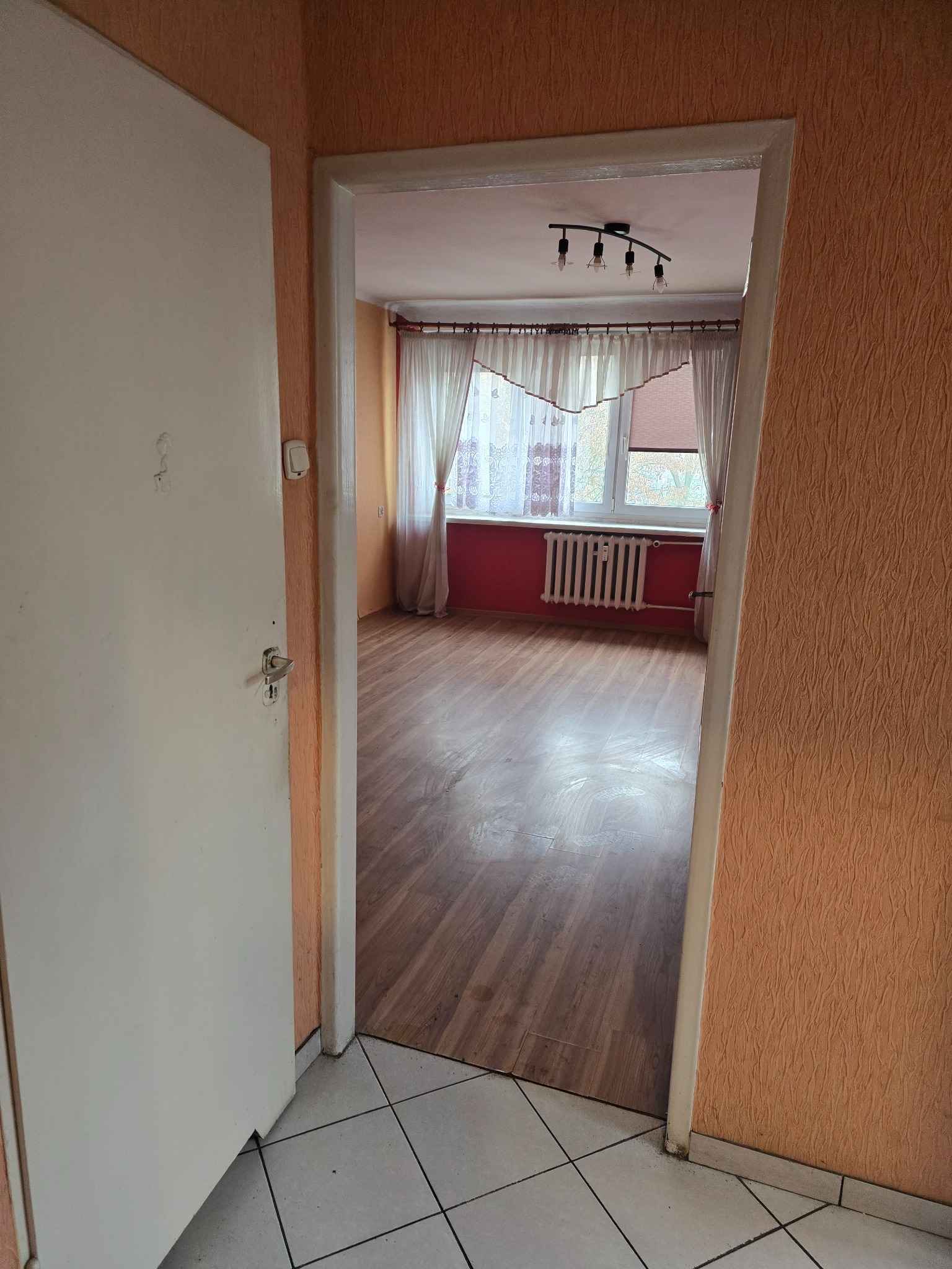 Pusty pokój z drewnianą podłogą widziany przez otwarte drzwi. Pomarańczowe ściany, białe zasłony i grzejnik pod oknem. Widoczny fragment korytarza z białą podłogą.