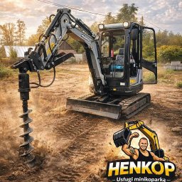 Henkop - usługi minikoparką Henryk Kubiciel - Mini koparka z wiertłem w akcji, logo firmy Henkop w prawym dolnym rogu. Ślady opon na ziemi, w tle budynek i drzewa. Słoneczny dzień.