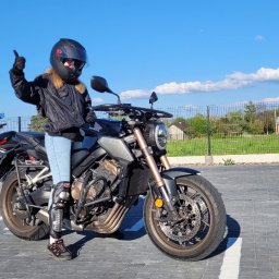 OSK POL-CAR Jan Ziemiański - Kobieta w kasku na motocyklu Honda CB650R, uniesiona ręka w geście aprobaty. W tle zaparkowane motocykle i samochód, błękitne niebo.