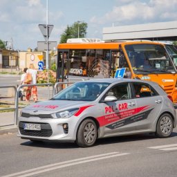 OSK POL-CAR Jan Ziemiański - Srebrny samochód z oznaczeniem 'L' i logo 'POL-CAR' na tle pomarańczowego autobusu. Pojazd szkoły jazdy na tle miejskiego krajobrazu w słoneczny dzień.
