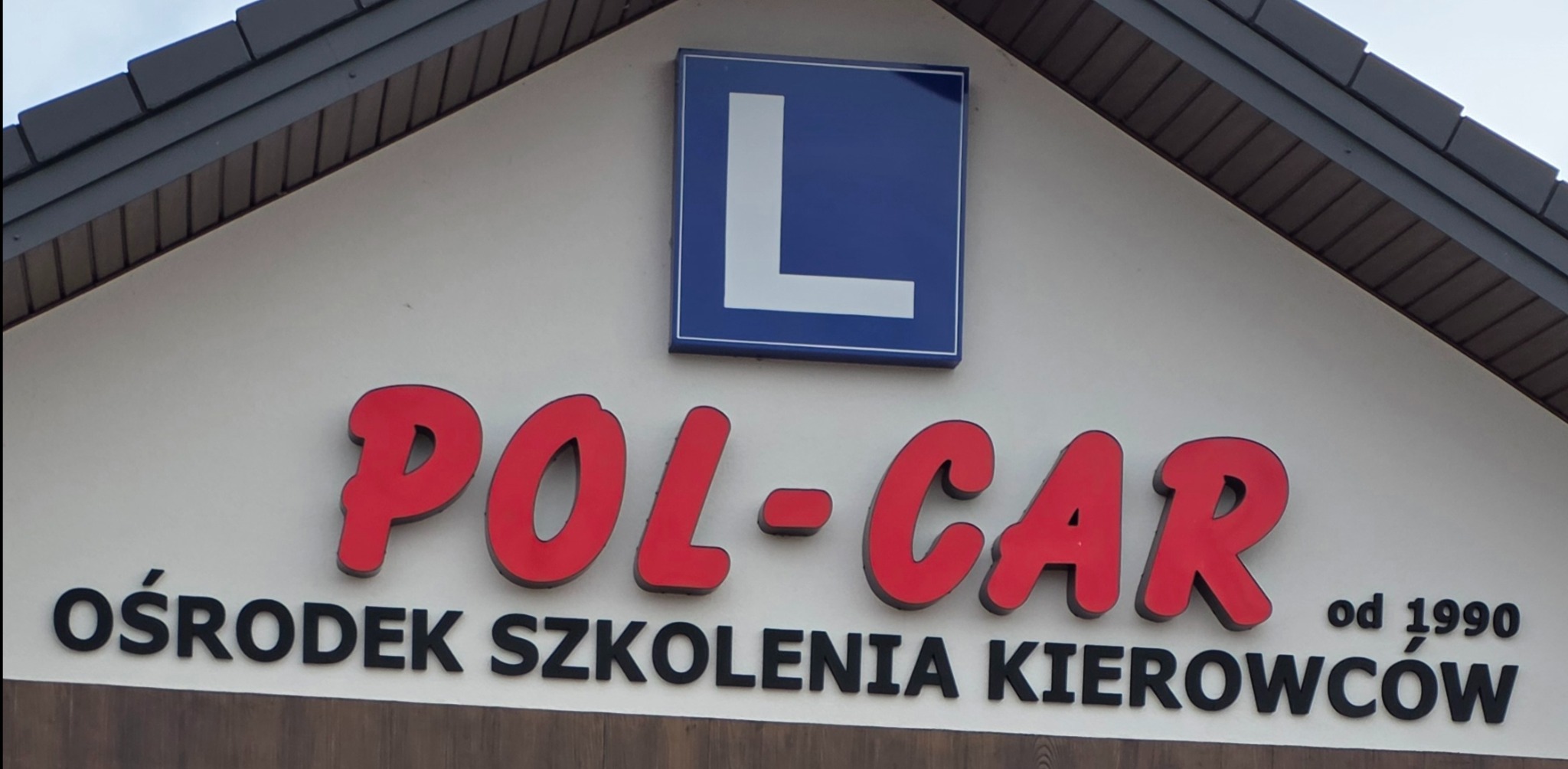 Fasada budynku z szyldem 'POL-CAR Ośrodek Szkolenia Kierowców od 1990' i symbolem 'L' szkoły jazdy. Czerwone litery na jasnym tle.