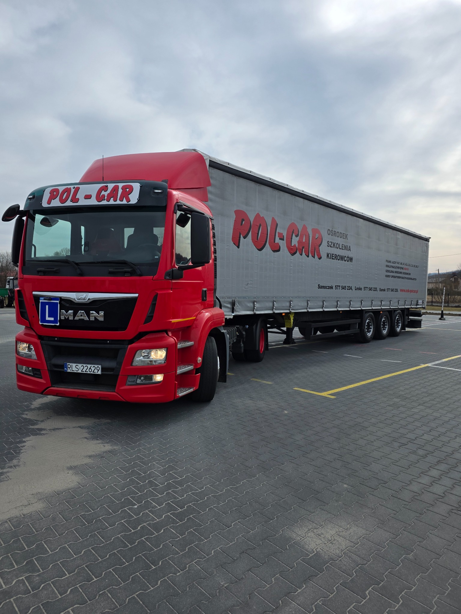 Czerwona ciężarówka MAN z przyczepą, oznakowana logo 'POL-CAR' i literą 'L', na placu manewrowym ośrodka szkolenia kierowców. Widoczne numery kontaktowe.
