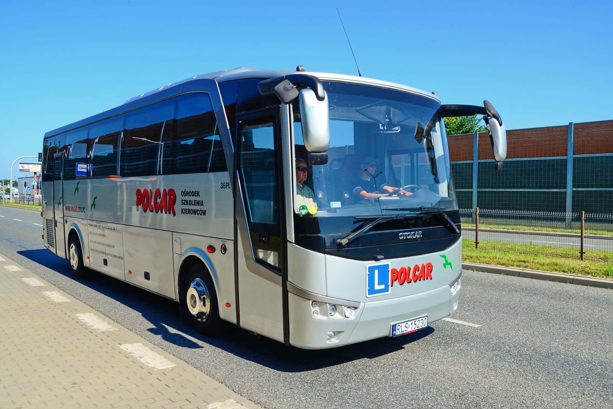 Autobus szkoleniowy Polcar z oznaczeniem 'L' na przodzie, jadący po drodze w słoneczny dzień. Widoczni instruktor i kursant za kierownicą.