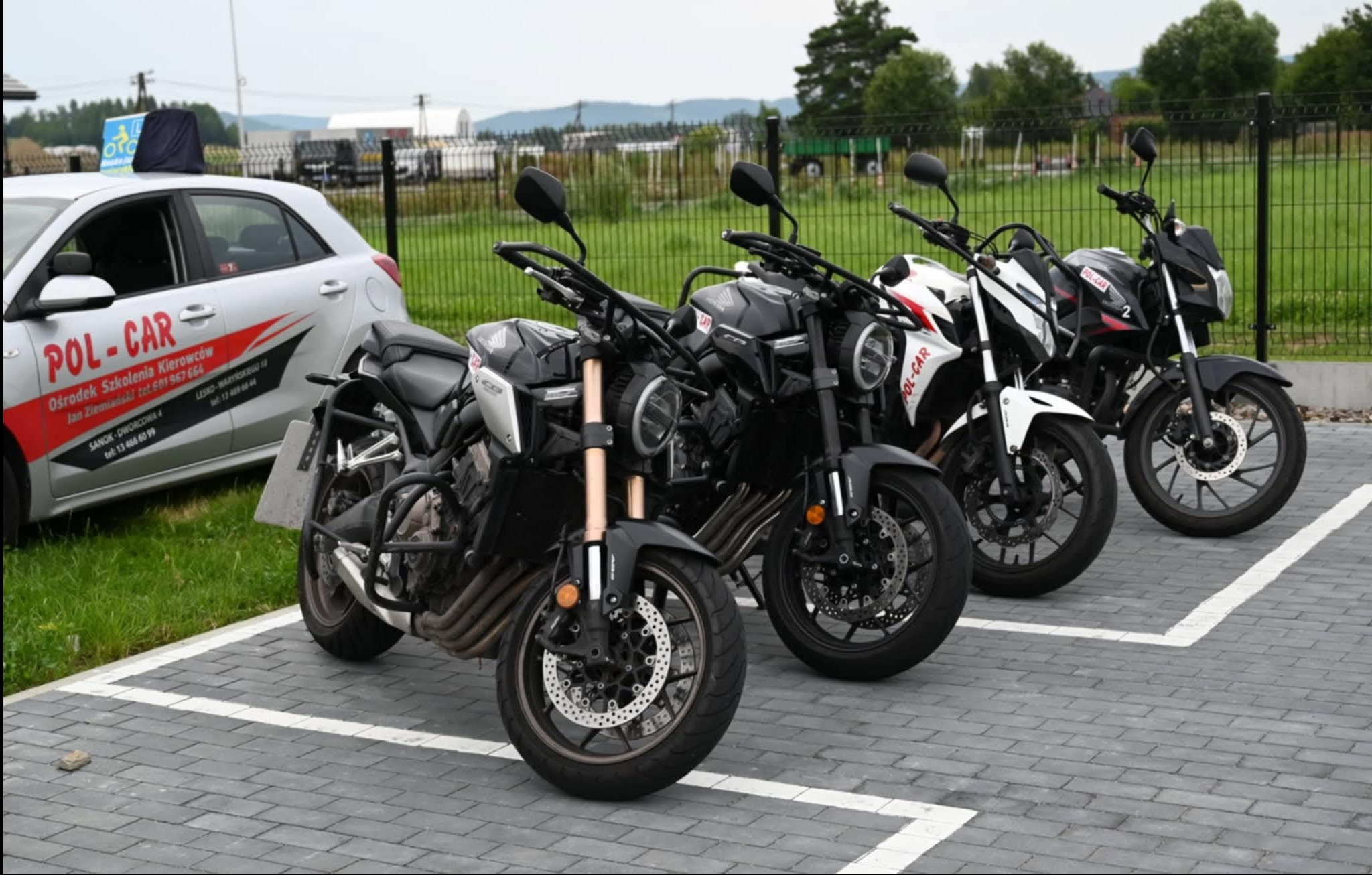 Cztery motocykle szkoły jazdy Pol-Car zaparkowane na szarym bruku, z samochodem nauki jazdy w tle i zielonym krajobrazem. Widoczne logo firmy na pojazdach.
