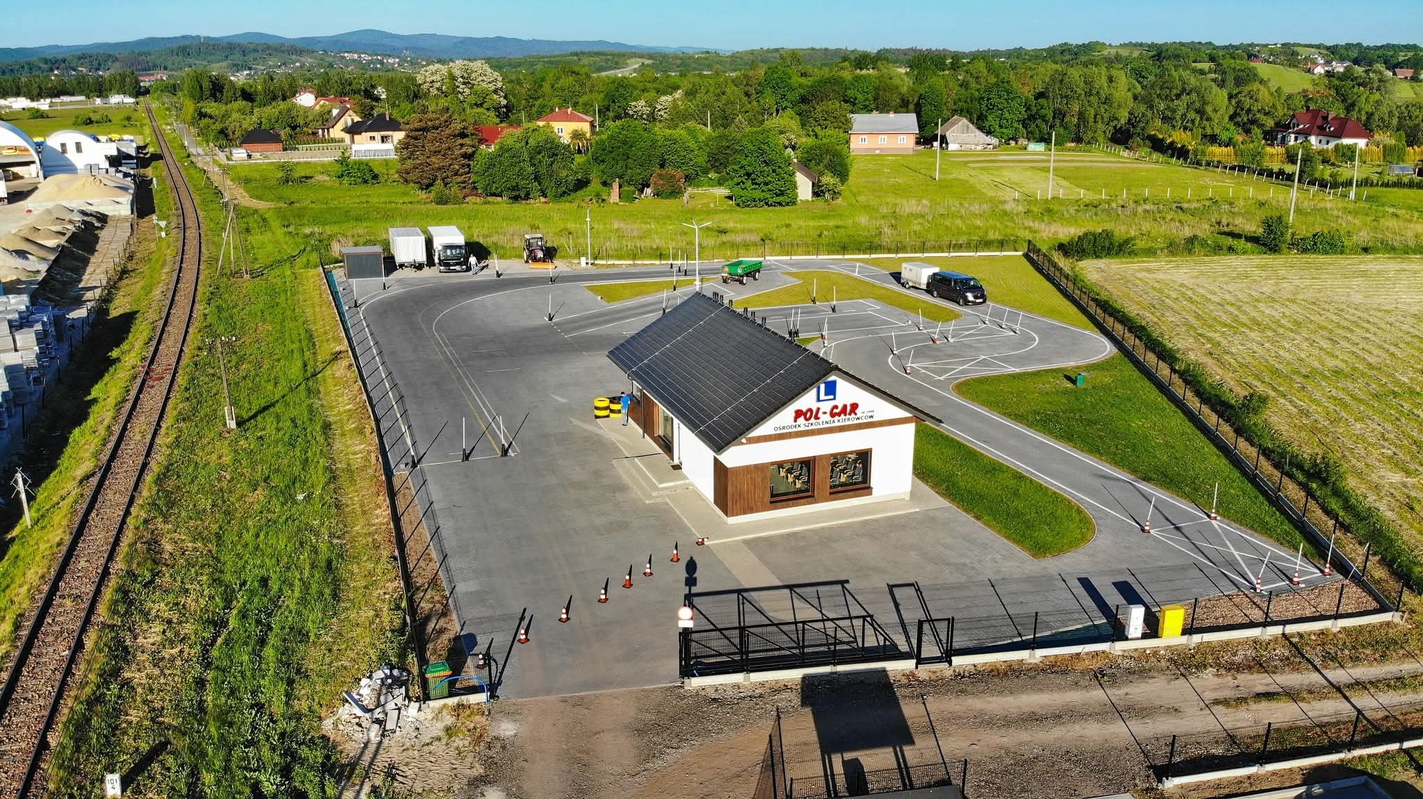 Widok z lotu ptaka na plac manewrowy szkoły jazdy z budynkiem biura, pojazdami i elementami infrastruktury szkoleniowej w otoczeniu zieleni i zabudowań.